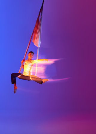 Strong man, professional aerial gymnast, acrobat training with aerial tissues against gradient blue purple studio background in neon with mixed lightsの写真素材