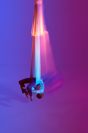 Young male professional aerial gymnast, acrobat training on aerial fabric against gradient blue purple studio background in neon with mixed lightsの写真素材