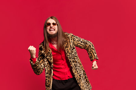 Portrait of young man with long hair posing in stylish fur coat and sunglasses against red studio background. Emotionally shoutingの写真素材