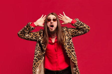 Fashion diva. Portrait of young man with long hair posing in stylish fur coat and sunglasses against red studio backgroundの写真素材