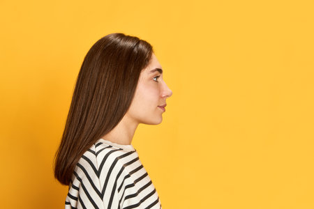 Side view portrait of young girl with brown straight hair, in striped sweater posing, looking away agaisnt yellow studio backgroundの写真素材
