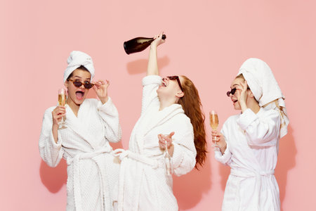 Three happy, beautiful girls in bathrobes drinking champagne, having celebration and fun against pink studio backgroundの写真素材
