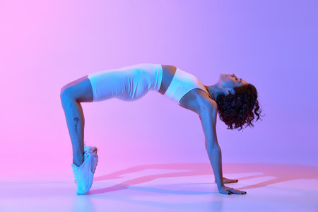 Young woman with strong, slim, fit body in white sportswear training against gradient pink blue background in neon lightの写真素材