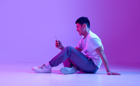 Happy, smiling young man in casual clothes sitting on floor, looking on mobile phone, texting...