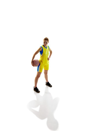 Full-length image of young girl, female basketball player in yellow uniform posing with ball against white studio background. Isometric view. Concept of professional sportの写真素材