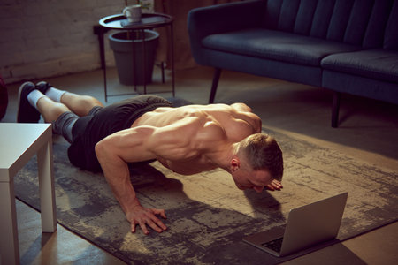 Sportive young man with strong, muscular body training shirtless, doing push-up exercises at home with online lessons on laptopの写真素材