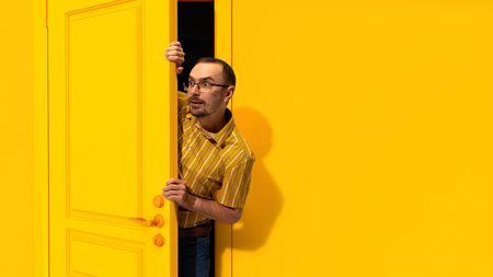 Young man in glasses peeking out yellow door and looking with excitement, shock and positive impression. Big sales, success, winの写真素材