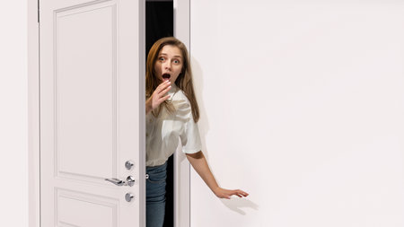 Young woman peeking out open grey door, covering mouth with hand. Looking shocked, surprised, astonished. Unexpected situationの写真素材