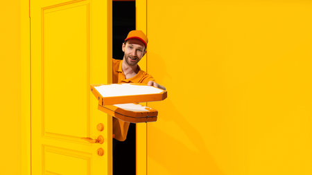 Young delivery man in orange uniform peeking out yellow door with pizza boxes. Fast and convenient delivery service. Online shoppingの写真素材