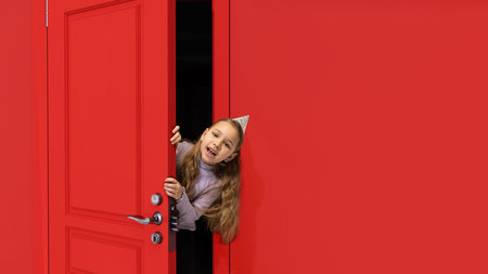 Cute, beautiful little girl, kid in birthday accessories peeking out red door with surprised face. Cheerful celebrationの写真素材