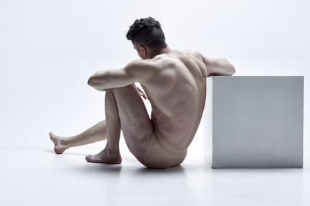 Man with relief, muscular, strong body shape sitting, posing shirtless in underwear against white studio background. Back viewの写真素材