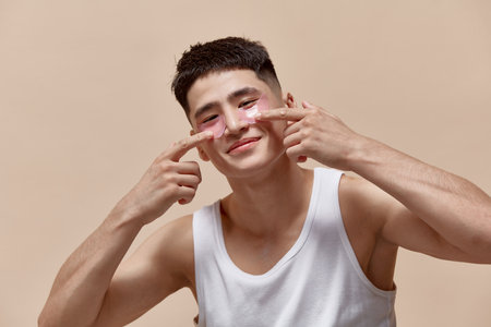 Portrait of handsome young asian guy taking care after skin, applying under eye patches against light brown studio backgroundの写真素材