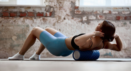Young girl with slim, sportive body training, doing body, back massage with foam roller on mat indoors on daytimeの写真素材