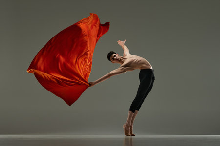 Graceful, handsome, young man, professional ballet dancer dancing with red silk fabric against grey studio background.の写真素材