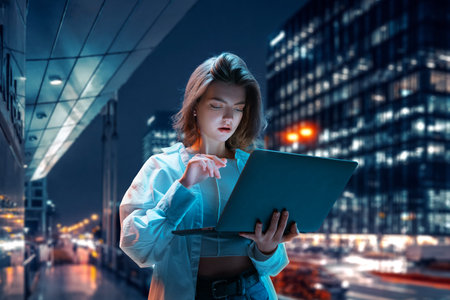Pretty young girl standing on street in evening and working on laptop. Freelance job and education. Blurred city backgroundの写真素材