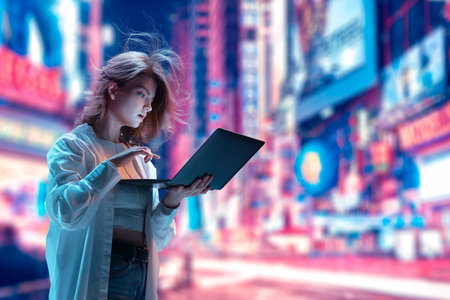 Beautiful young girl standing on lively street and looking on laptop. Working, studying, shopping. Unfocused city backgroundの写真素材