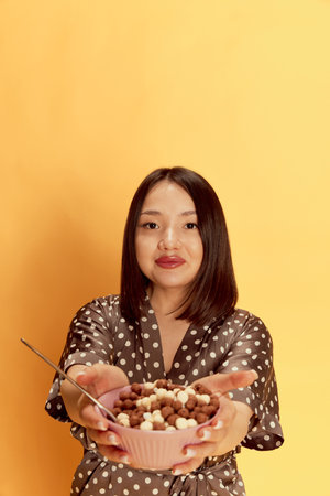 Portrait of young asian girl in comfortable homewear, holding bowl with cereal against yellow studio background. Morning, breakfastの写真素材