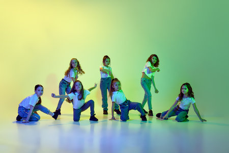 Group of little girls, children in casual clothes dancing, learning modern dance styles against green studio background in neon lightの写真素材