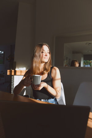 Portrait of young beautiful woman spending cozy time at home on day with sunlight. Sitting at table and drinking morning coffeeの写真素材