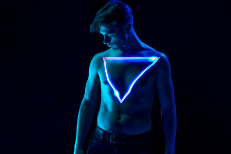 Handsome, muscular, young man posing over dark background with digital neon triangle reflection on body. Masculinityの写真素材