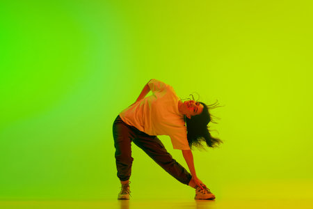 Artistic, flexible, positive young girl, hip-hop dancer performing against gradient green yellow background in neon lightの写真素材