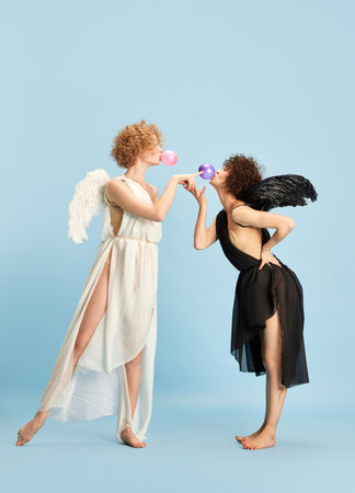 Tandem of evil and kindness. Portrait of two beautiful young women in images of angel and demon against blue backgroundの写真素材