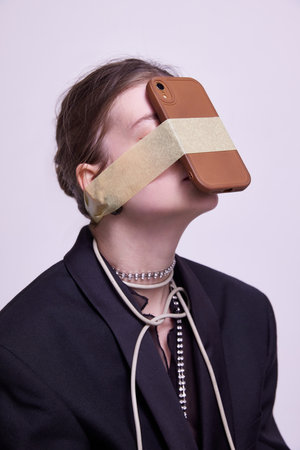 Social media addiction. Stylish young girl sitting with phone sticking to her head over grey studio backgroundの写真素材