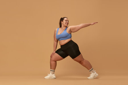 Young smiling woman with fat, overweight body training in sportswear against brown studio...