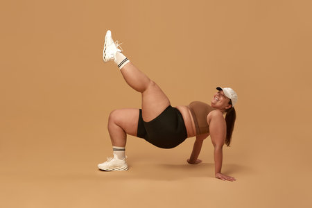 Positive, young, overweight woman training in stylish and comfortable sportswear against brown studio background. Full-body workoutの写真素材