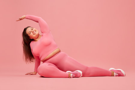 Well-being and self-love. Young overweight woman training in pink sportswear, doing stretching against pink studio backgroundの写真素材