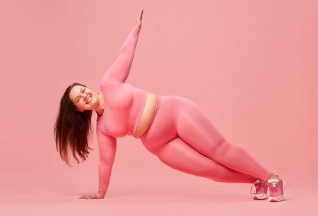 Young, positive, motivated woman with overweight body training in sportswear against pink studio background. Exercisingの写真素材