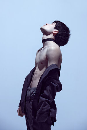 Muscular body. Handsome, stylish male model, young guy posing shirtless in black jacket and pants against light blue studio backgroundの写真素材