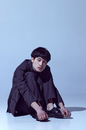 Classical look. Portrait of handsome young guy posing in black jacket, tie and pants against light...