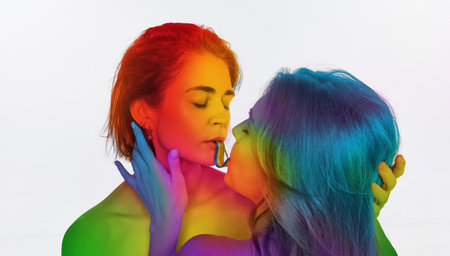 Portrait of loving young women kissing, eating jelly candy against white background. Models painted into rainbow colors. Lgbtの写真素材