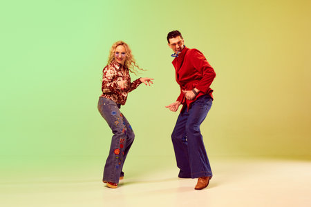 Young emotional couple, man and woman in stylish retro clothes dancing against gradient green yellow...