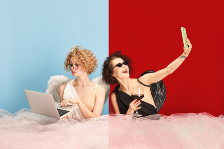 Young woman in image of angel and demon sitting at table and using gadgets against blue red background. Work and bloggingの写真素材