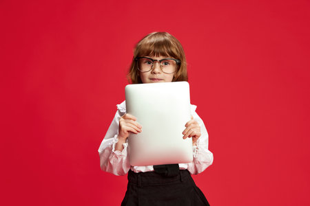 Portrait of cute little girl, child in big glasses, holding tablet against red studio background....