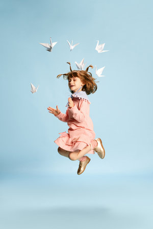 Full-length portrait of playful, lovely little girl in pink dress posing, playing with paper birds against blue studio backgroundの写真素材