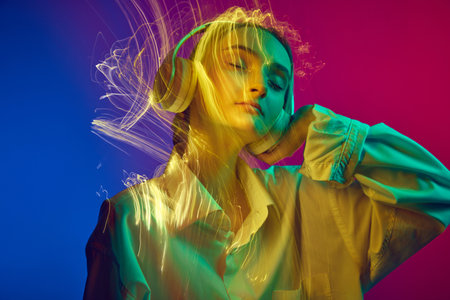 Portrait of young girl listening to music in headphones, posing over gradient blue pink background...