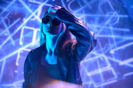 Night life. Urban fashion. Portrait of young beautiful girl in leather jacket posing over blue background with abstract neon elements.の写真素材