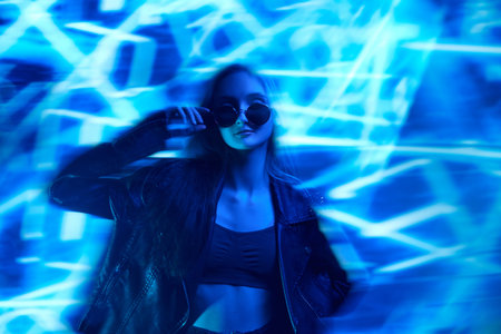 Unfocused portrait of young beautiful girl in leather jacket posing over blue background with abstract neon elements.の写真素材