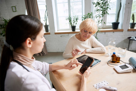 Senior woman and female doctor using gadgets to check patients health information. In office. Modern...
