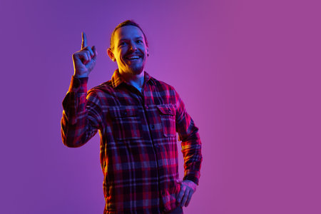 Portrait of thoughtful man in checkered shirt generating creative ideas, raising finger up against purple studio background in neon lightの写真素材