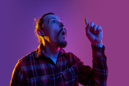 Portrait of thoughtful man in checkered shirt generating creative ideas, raising finger up against purple studio background in neon lightの写真素材