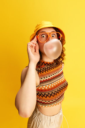 Portrait of funny, emotive young girl in panama and glasses, blowing bubble gum against yellow studio backgroundの写真素材