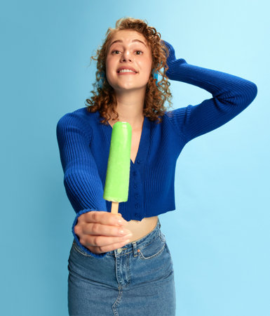 Portrait of young girl in blue top and jeans skirt, listening to music in headphones and giving ice cream against blue studio backgroundの写真素材