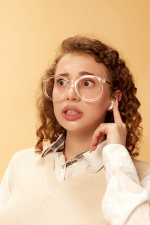 Portrait of young curly girl in white blouse and glasses, pressing on headphones, talking on phone studio background. Business womanの写真素材