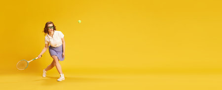 Portrait of young, concentrated girl in sportswear, tennis playing in motion hitting ball with racket against yellow studio backgroundの写真素材