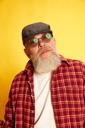 Portrait of stylish, mature, bearded man in checkered shirt, glasses and cap posing against yellow studio backgroundの写真素材