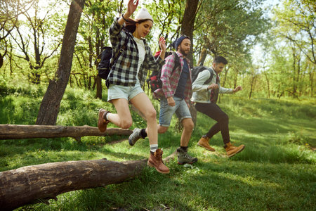 Group of young people, friends walking in forest, going hiking on warm sunny day, jumping over big...